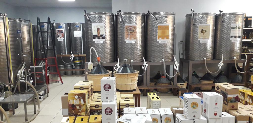 Serbatoi in acciaio inox con etichette, damigiane in cesti di vimini e scatole bag-in-box da 5 litri impilate sul pavimento
